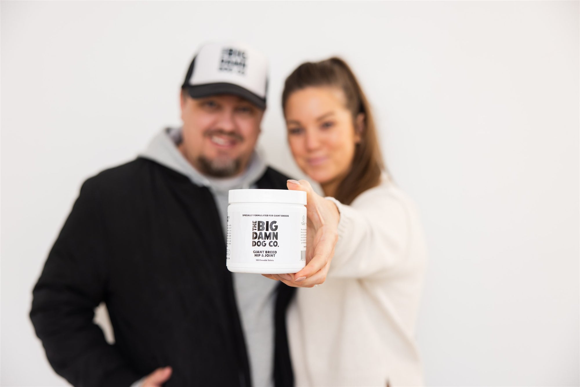 A man and a woman hold up a white plastic tub of Big Damn dog Co. joint care toward the camera against a plain white background.