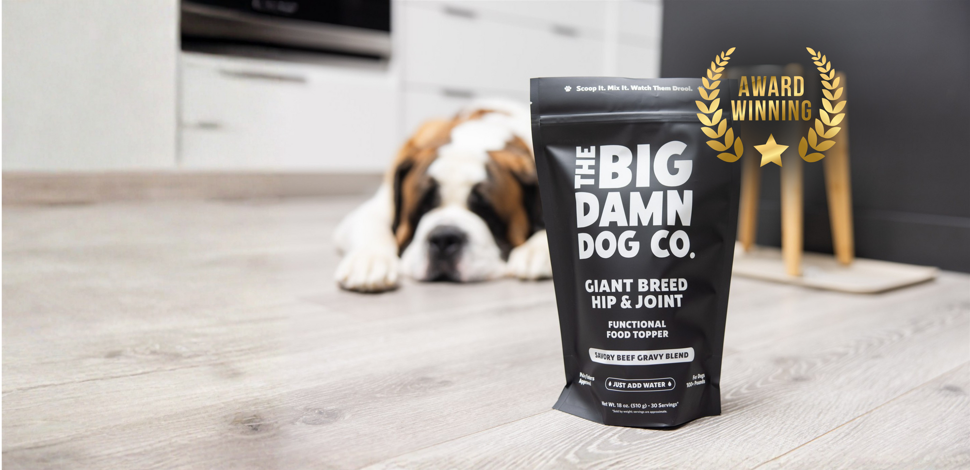 A black stand-up pouch of The Big Damn Dog Co. Giant Breed Hip & Joint functional food supplement stands on a light wood floor in a kitchen setting, with a Saint Bernard dog lying blurred in the background.