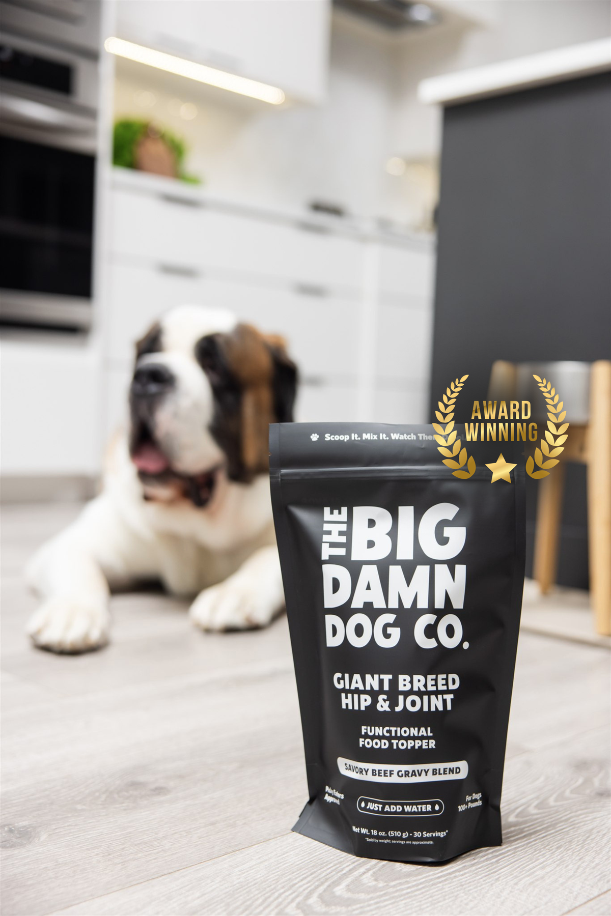 A black stand-up pouch of The Big Damn Dog Co. Giant Breed Hip & Joint supplement is shown in the foreground on a light wood floor, with a Saint Bernard dog lying blurred in the background in a modern kitchen setting.